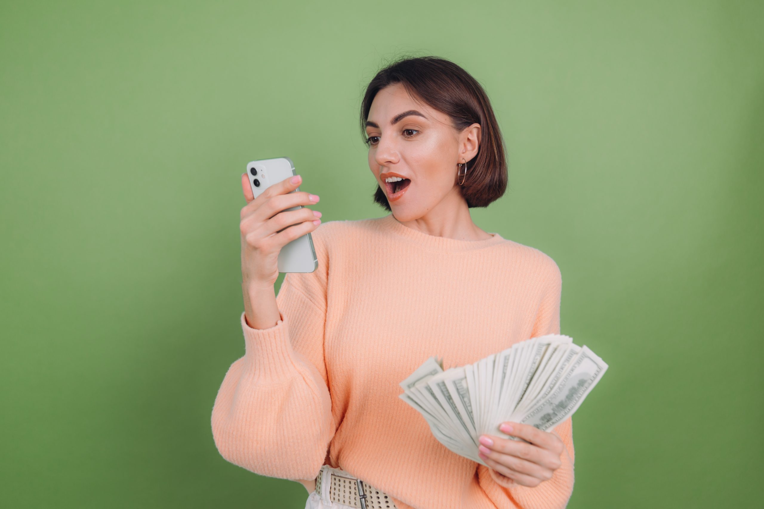 Young woman in casual peach sweater  isolated on green olive background excited holding black screen phone and money amazed surprised winner  emotions