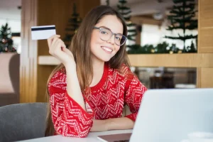 MULHER FELIZ COM CARTÃO DE CRÉDITO SEM ANUIDADE USANDO COMPUTADOR E LENDO DICAS SOBRE COMO ESCOLHER O MELHOR CARTÃO DE CRÉDITO SEM ANUIDADE.
